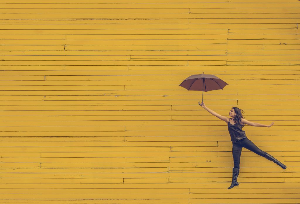 woman jumping in the air with a black umbrella against a yellow wall